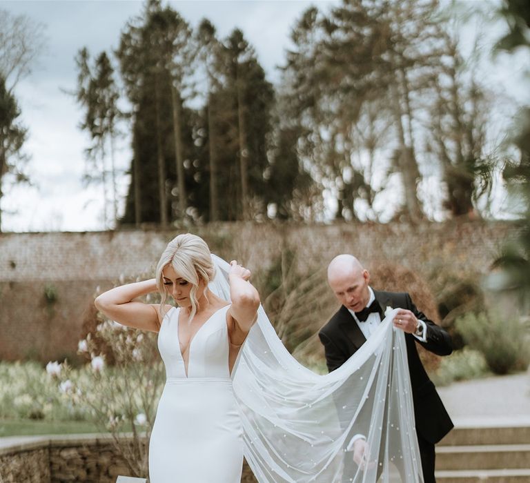 Groom in black tie helps the bride with her pearl veil with the bride wearing a fitted made With Love wedding dress 