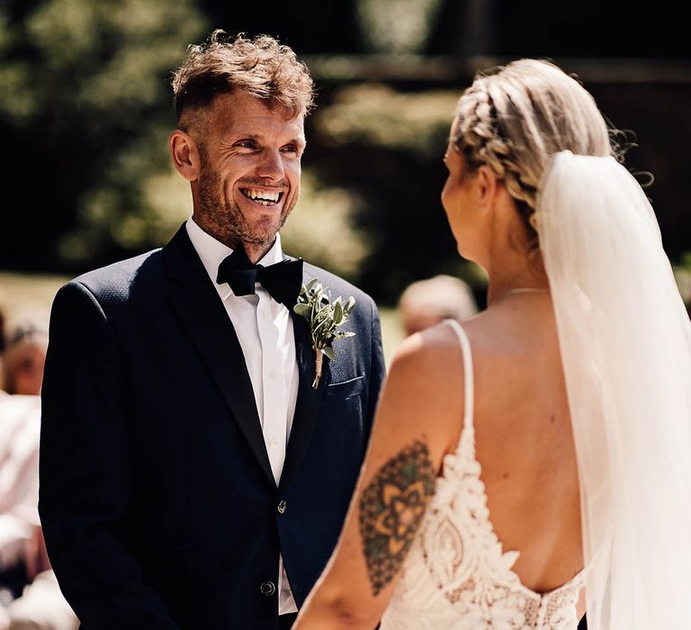 Groom in blue tuxedo and black bow tie smiles as he looks at the bride and they hold hands together for outdoor ceremony 