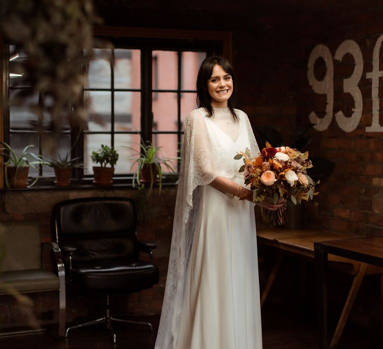 Bride holds Autumnal floral bouquet and wears lace cape whilst stood in The Chimney House on her wedding day