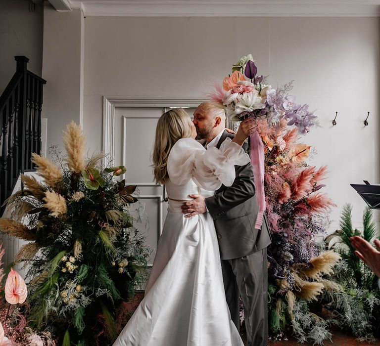 Groom kissing his bride in a JESUS PEIRO wedding dress at their wedding ceremony surrounded by colourful colour flower arrangements 