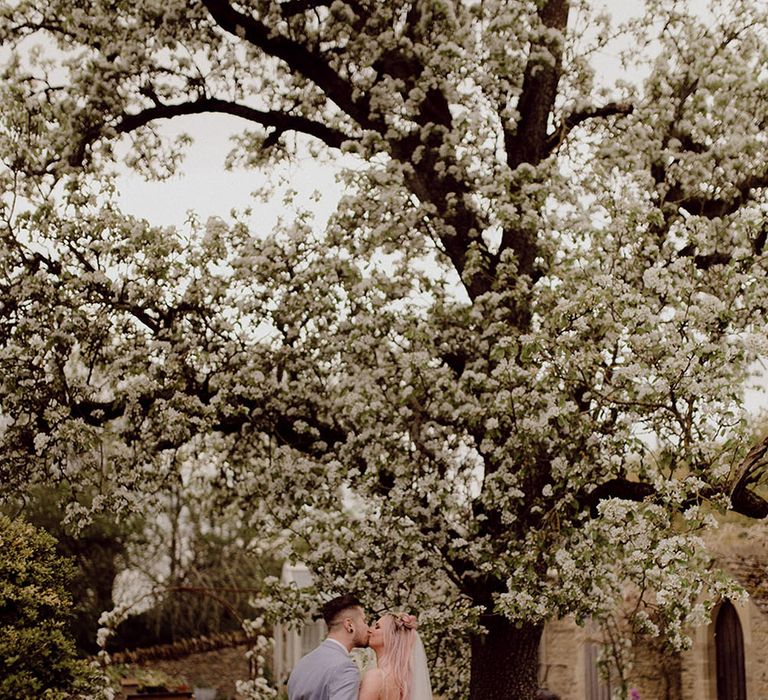 Bride & groom kiss beneath white blossom tree at Oxleaze Barn on their wedding day