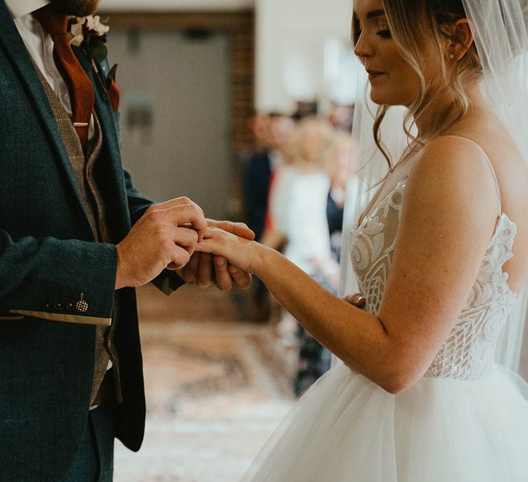 Groom in blue suit and brown waistcoat puts the ring on the bride's finger during their wedding ceremony 