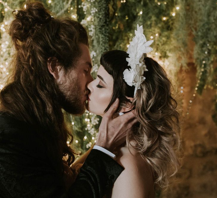 Groom with long hair kissing his bride in a white 3D flower headband at altar with a twig, fairy light and foliage backdrop 