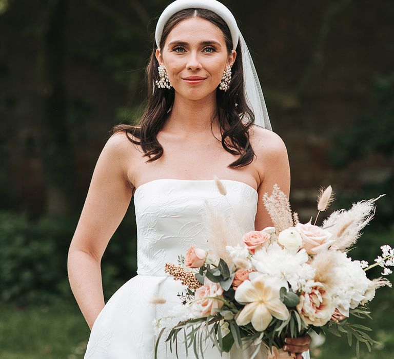 Smiling bride in brocade Jesus Peiro wedding dress with pearl earrings and headband carrying blush pink and dried grass bouquet 