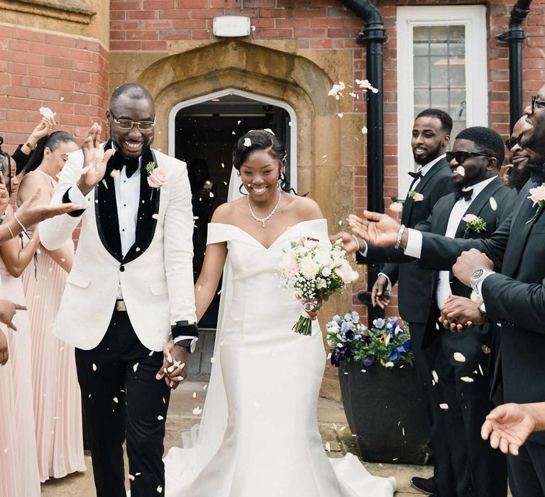 Bride wearing fitted off the shoulder wedding dress and carrying pastel pink and white bouquet walks with her groom in black tie through confetti thrown by wedding guests