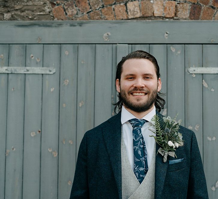 Groom wears three piece suit with blue jacket and grey waistcoat complete with oversize floral buttonhole and floral styled tie