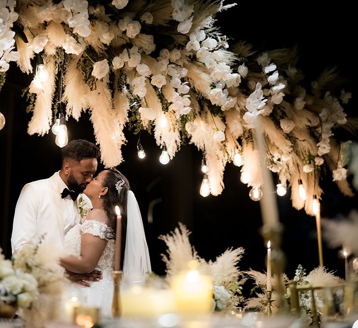 Pampas grass installations with festoon lighting hang above bride & groom as they kiss 