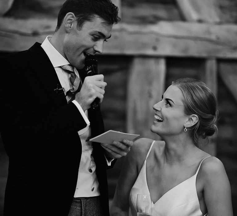 Groom in a morning suit reads out his speech as the bride listens and looks up at him 