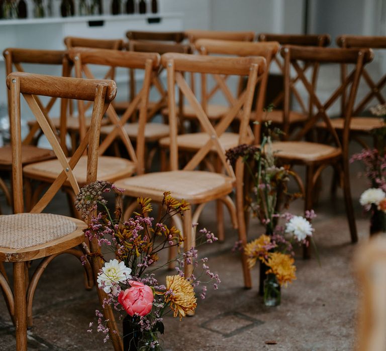 Small floral arrangements in bright colours beside wooden chairs lining the aisle