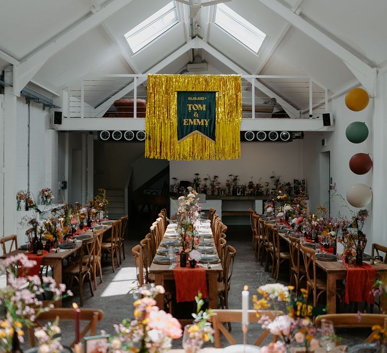 Industrial styled wedding decor complete with gold streamers and wooden banquet tables with orange fabric table runner and small floral arrangements