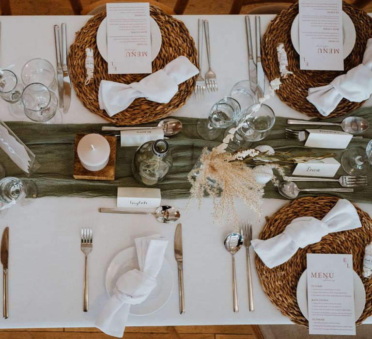 Boho, rustic and earthy tablescape design with bottle green table runner, wicker place mats, white napkins, pillar candle, and dried flower arrangements in small glass vases and bottles