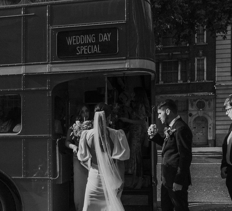 Bride with open back puff sleeve wedding dress and pearl detail veil with groom getting on bus wedding transport