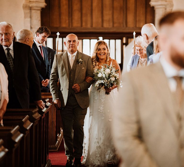 Father of the bride in a dark grey suit walks the bride in a lace wedding dress down the aisle to the groom waiting 