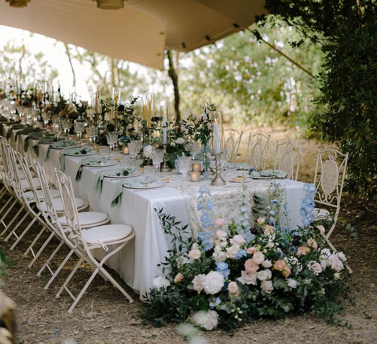 Beautiful Italian tablescape with ribbed glassware, gold candlestick holders and flower arrangements that include thistles, peonies, carnations and wildflowers under a marquee with rattan lamps