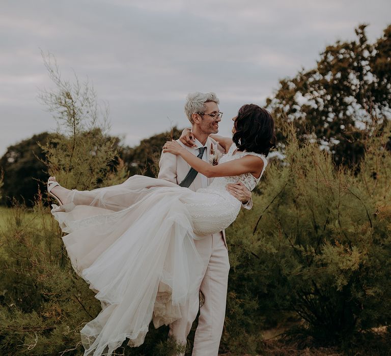 Groom in light pink suit carries bride wearing lace and tulle fishtail dress in front of greenery