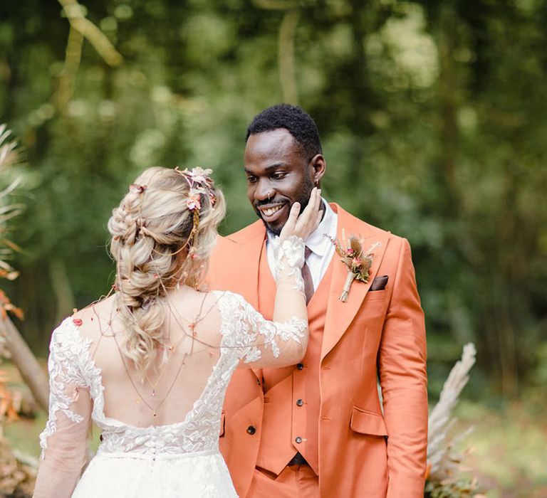 Bride in open back detail boho wedding dress with botanical embroidered details and headpiece with delicate details and mix of soft gold & bronze metals embracing groom in burnt orange three piece wedding suit with dried flower boutonniere and brown pocket square