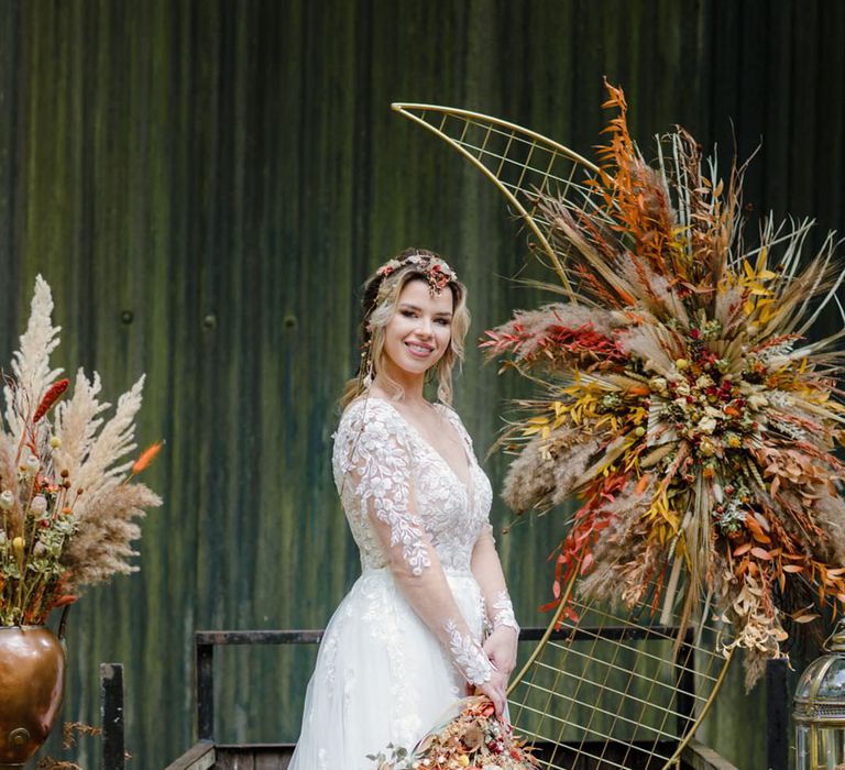 Bride standing by giant gold crescent moon ceremony backdrop on back of a wooden trailer wearing long sleeved v neck soft ivory dress with lace floral detailing on the bodice and sheer sleeves with autumnal dried flower arrangements