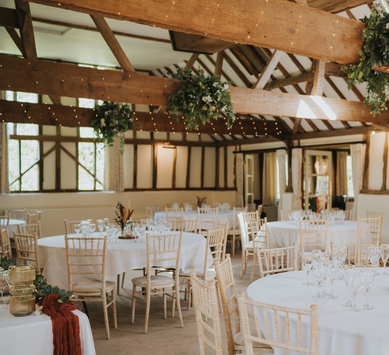 Reception room at Essex wedding venue with white neutral tablescapes, large foliage and white rose floral arrangements and fairy lights in the beams of the ceiling 