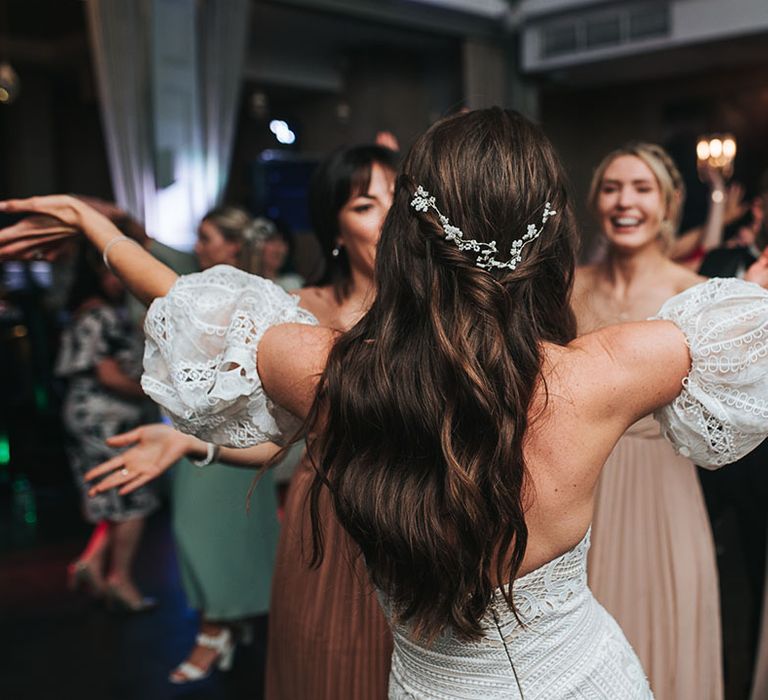 Bride wears her hair in a half up half down style with white flower accessory in a strapless lace gown with detachable sleeves dancing on the dance floor 