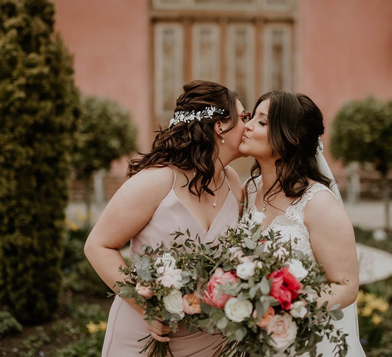 Bride in lace wedding dress kisses her bridesmaid in pale pink bridesmaid dress