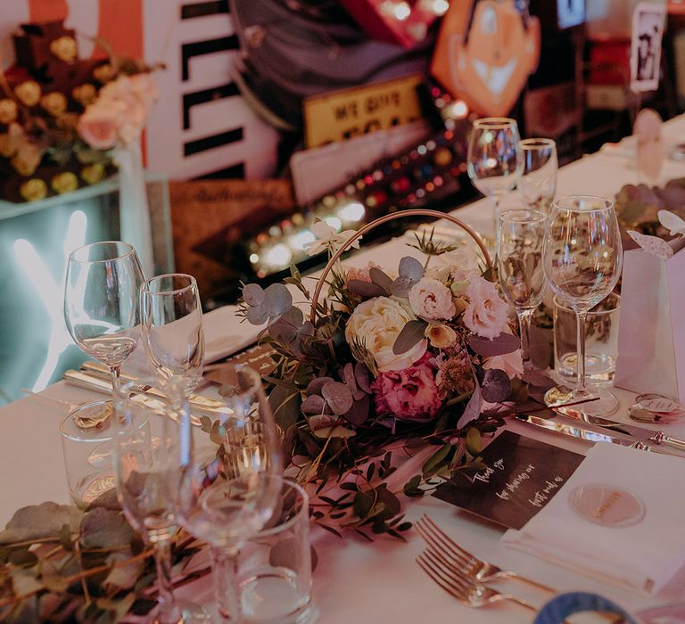 Rustic wedding tablescape with foliage table runners and rose and peony centrepieces with colourful neon signs in the background 