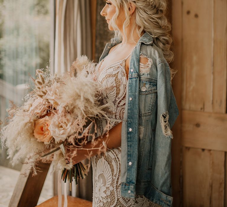 Bride in a boho lace wedding dress with a ripped light blue wash denim jacket with platinum blonde hair in a fishtail braid hairstyle 