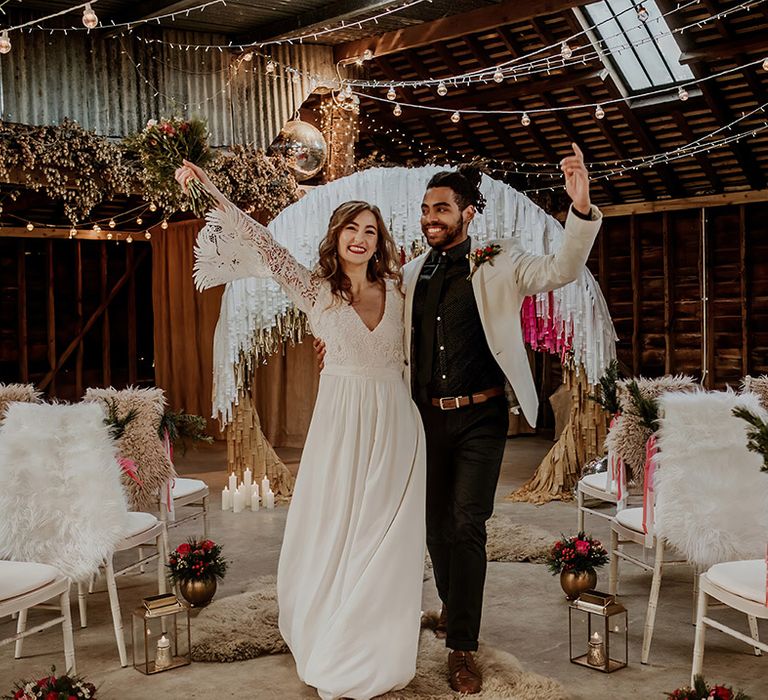 Bride in v-neck boho long sleeve lace wedding dress with tassels and groom in white blazer with colourful boutonniere, black shirt and black suit trousers cheering walking down the aisle of barn wedding venue 