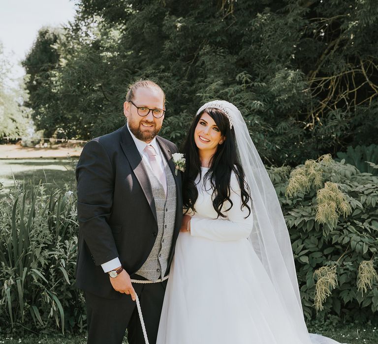 Groom in dark grey and light grey suit holding their pet dog, Stitch on a lead with the bride in a long sleeve high neck wedding dress and veil 