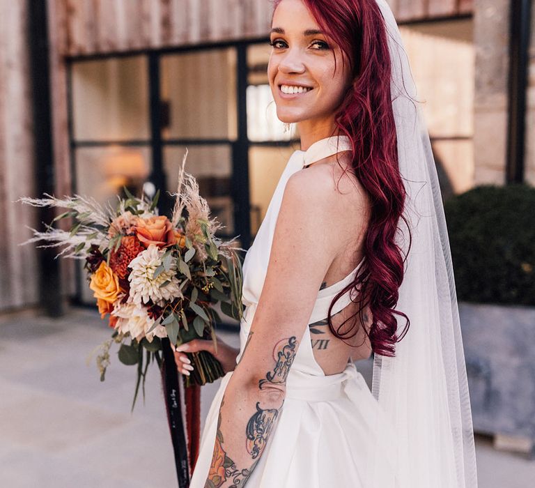 Bride holding orange autumnal wedding bouquet for September wedding with her red hair curled wearing a Halfpenny London wedding dress 