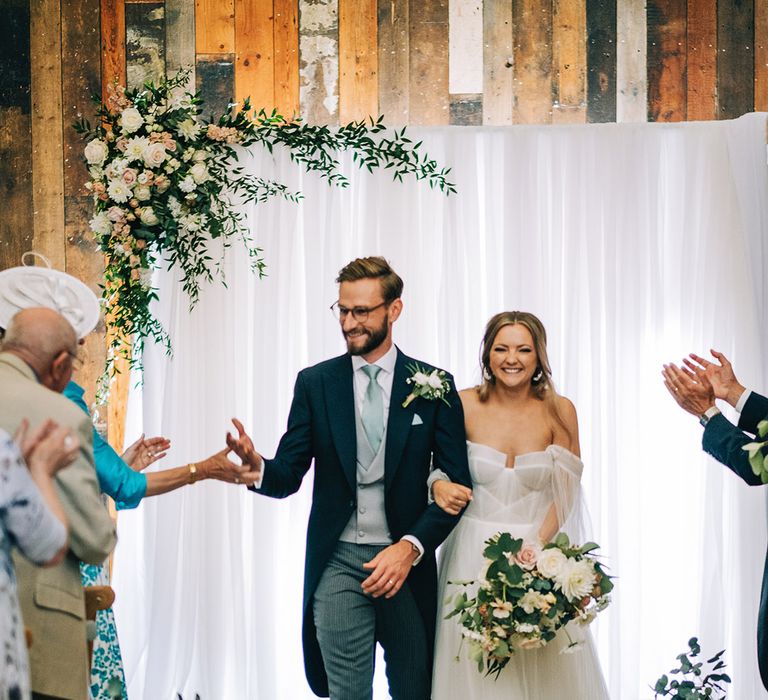 Groom in dark blue morning suit jacket with grey pinstriped trousers, light blue tie and waistcoat walking with the bride in long sleeve whimsical wedding dress 