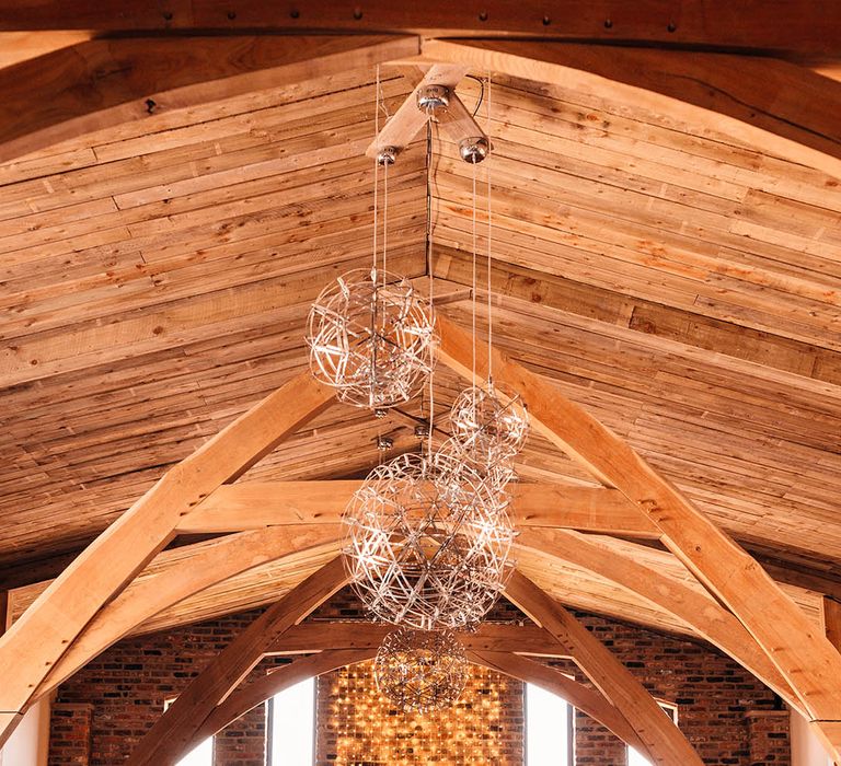 Brides stand holding hands for their wedding ceremony in front of column of fairy lights lighting up the cosy barn 