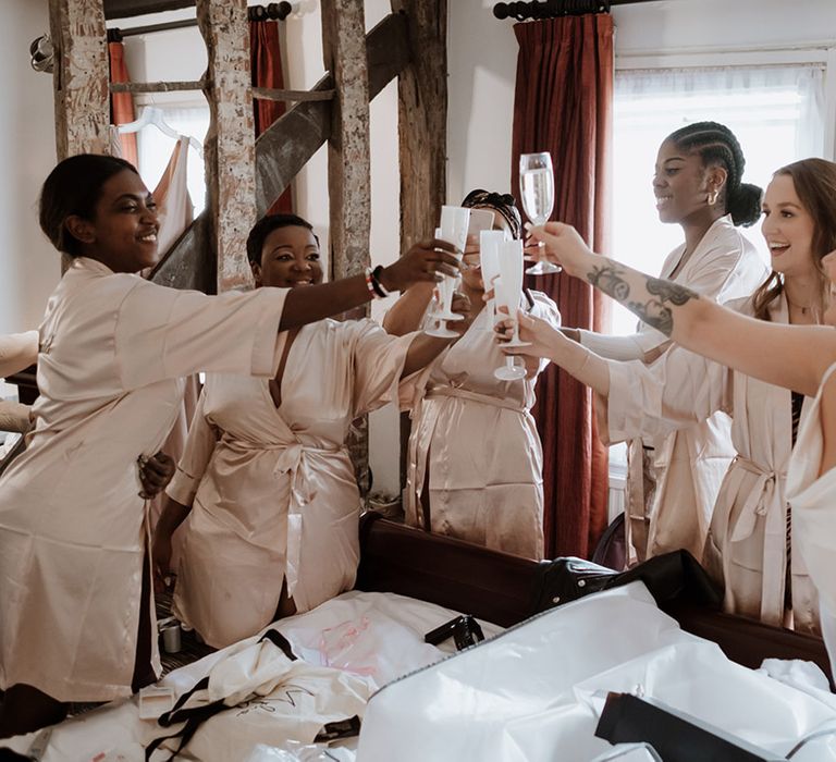 Bridesmaids in blush pink robes drinking from white champagne flutes with the bride to celebrate as they get ready together 