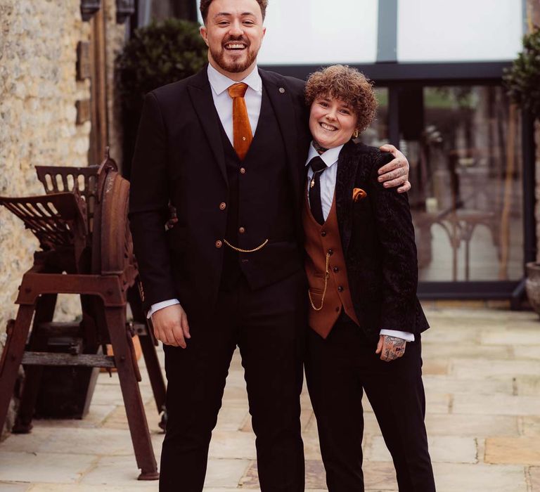 Bride in dark velvet bridal jacket with rust waistcoat and pocket square, black suit trousers, platform Dr Martens and gold pocket watch standing with wedding guest in dark suit with rust orange tie at Kingscote Barn Tetbury