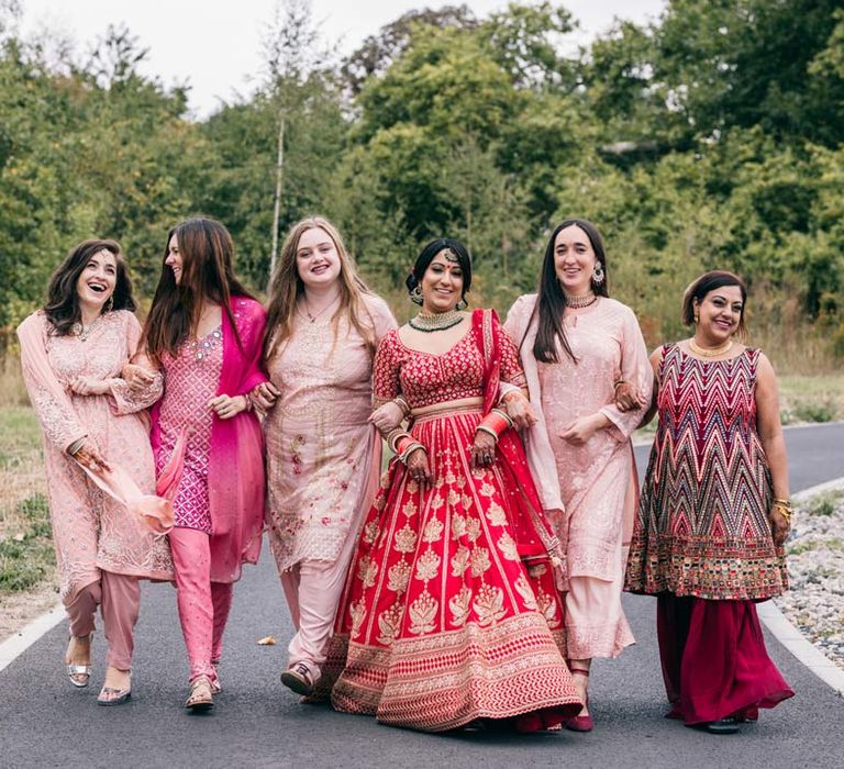 Bride in red and gold lehenga walking arm in arm with bridesmaids in traditional pink Indian bridesmaid outfits at Templars Barn