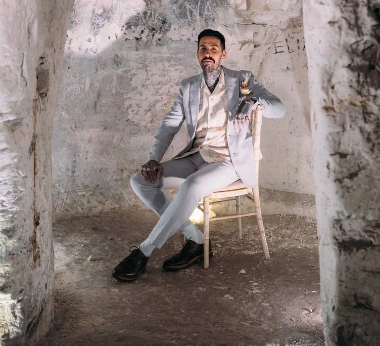 Groom in baby blue linen suit, personalised embroidered linen shirt and garden rose and pampas grass boutonniere sitting on chair in Margate Caves