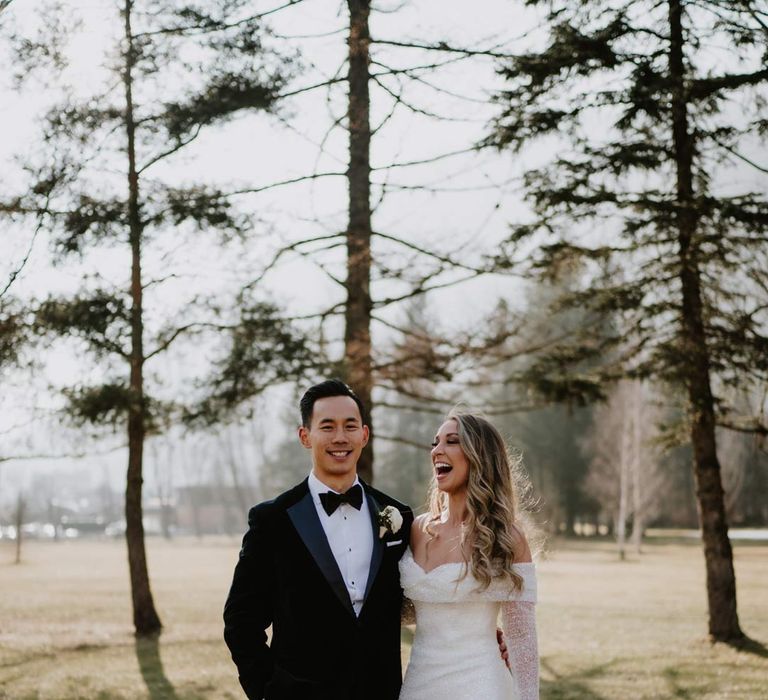 Bride in off shoulder sparkly mermaid wedding dress holding white garden rose, eucalyptus, baby's-breath and foliage bridal bouquet standing with groom in classic black tuxedo, black bowtie and white garden rose boutonniere at French destination ski wedding