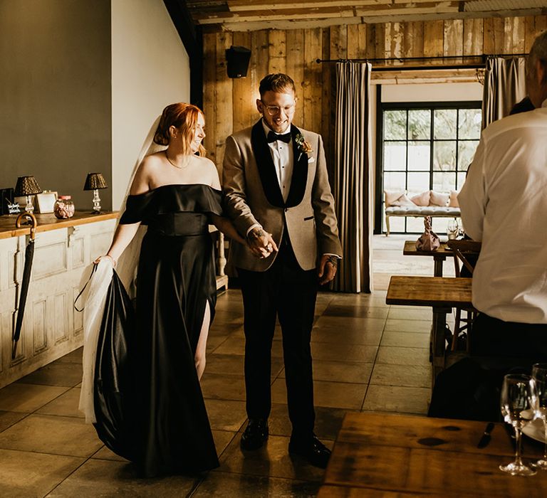 Bride in off the shoulder black wedding dress with groom in beige and black wedding suit entering their reception 