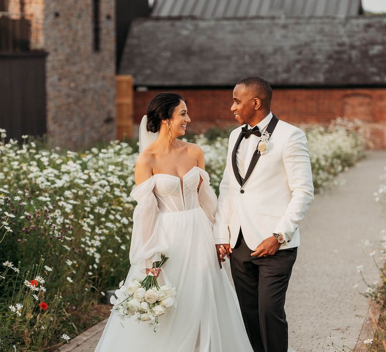 Bride in an off-the-shoulder wedding dress with front slit walks through daisy garden with groom in black-tie tuxedo