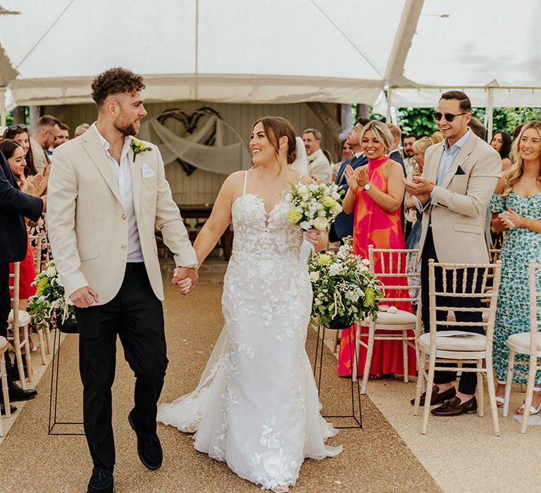 Bride in fitted lace wedding dress walking back down the aisle with the groom in a beige suit jacket 