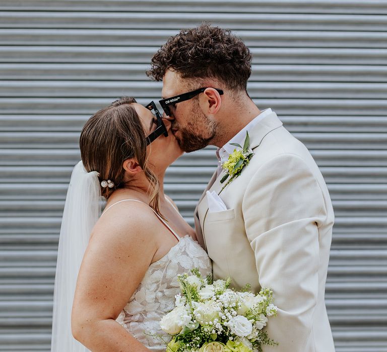 Bride wearing a lace wedding dress carrying a white flower bouquet kisses groom in beige suit at wedding 