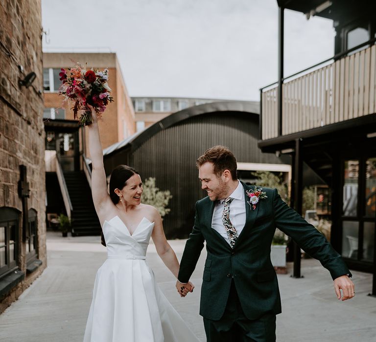 Bride in strapless Suzanne Neville wedding dress with front slit walking with groom in green suit 