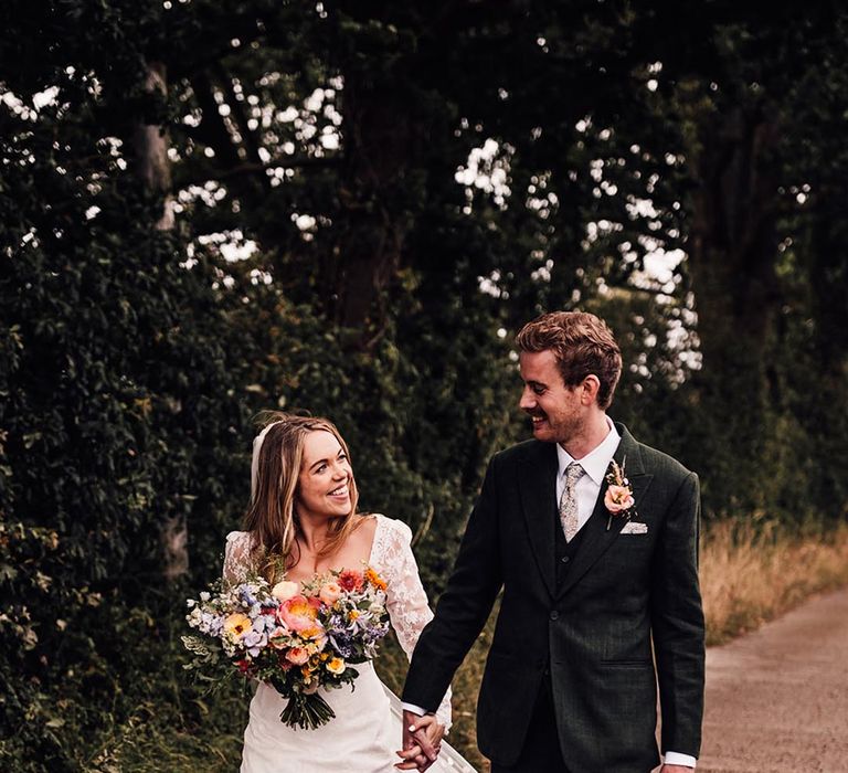 Bride in lace wedding dress holding colourful bouquet walking with the groom for couple portraits at rustic wedding 