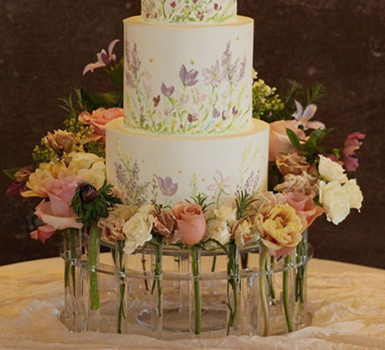 Three tier white iced wedding cake decorated with purple painted flowers surrounded by flower stems 