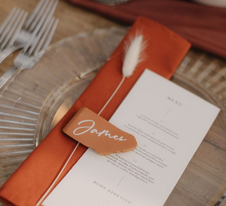 Rustic wedding tablescape with burnt orange napkin with wedding menu, name place name and bunny grass 