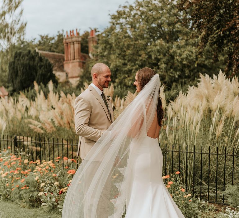 Bride wearing embroidered edge wedding veil looking at groom in beige wedding suit for cute couple portrait 