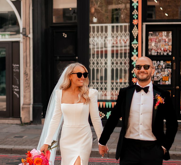 Bride wearing Suzanne Neville shoulder wedding dress with bright bouquet walking with groom in black tuxedo 