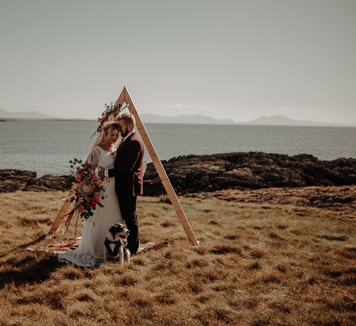 Bride and groom in front of boho wooden altar set up with dog in suit and large extravagant bouquet 