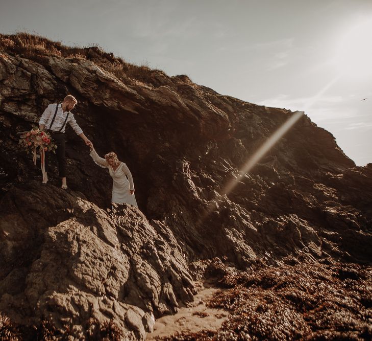 Groom helps bride climb the rocks in her lace tassel v-neck wedding dress