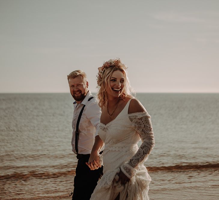 Bride smiles in flower crown and lace tassel wedding gown with layered necklaces and holds hands with groom in shirt and braces