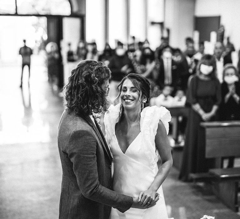 Bride in a ruffle sleeve wedding dress and groom with shoulder length curly hair laughing during their wedding ceremony 